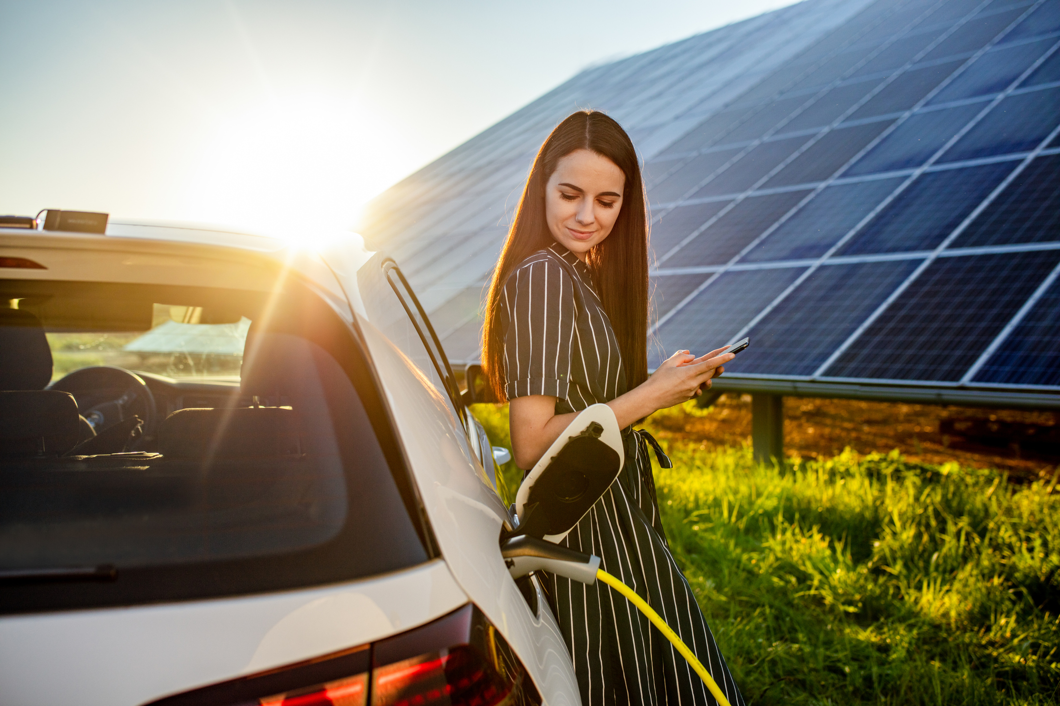 Waist up shot of a smiling young woman with black hair waiting for an electric car to charge and using a mobile phone with solar panels visible in the background